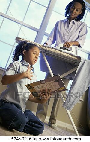 Stock Image - Mother ironing clothes with her daughter sitting on floor reading. Fotosearch