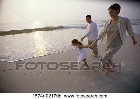 Stock Image - Parents running with their daughter on the beach. Fotosearch