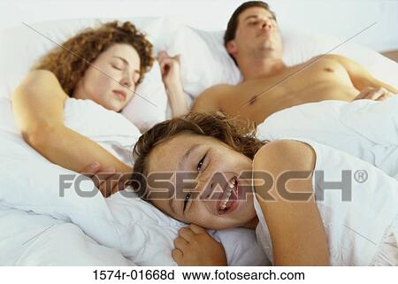 Portrait of a girl smiling with her parents sleeping on a bed behind her View Large Photo Image Stock Image - Portrait of a girl smiling with her parents sleeping on a bed behind her. Fotosearch