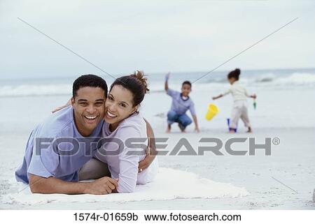 Stock Image - Portrait of parents lying on the beach with their son and daughter playing behind them. Fotosearch