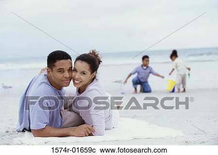 Stock Image - Portrait of parents lying on the beach with their son and daughter playing behind them. Fotosearch