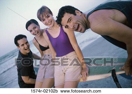 Stock Image - Portrait of two young men carrying two young women on a surfboard. Fotosearch