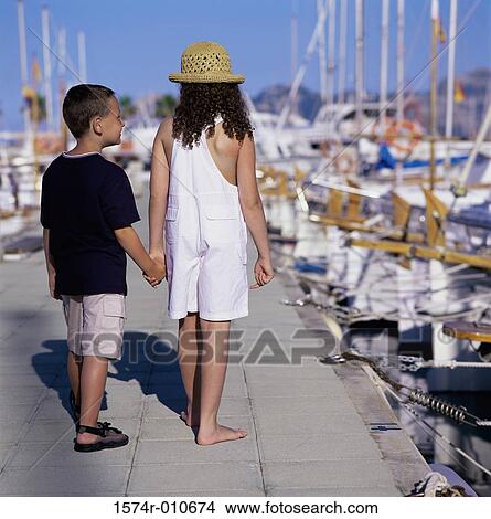 Picture - Rear view of a girl and a boy holding hands walking on a pier. Fotosearch