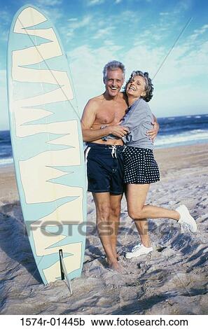 Stock Image - Senior couple standing on the beach near a surfboard. Fotosearch