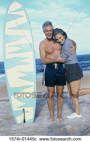 Stock Image - Senior couple standing on the beach near a surfboard. Fotosearch