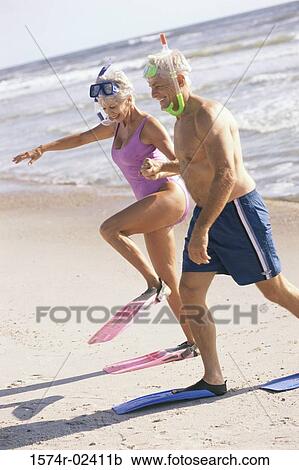 Stock Image - Senior couple walking on the beach wearing snorkeling gear. Fotosearch