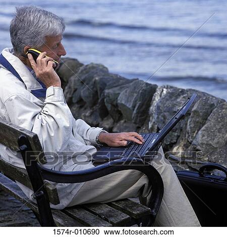 Senior man sitting on a bench using a laptop talking on a mobile phone View Large Photo Image Stock Image - Senior man sitting on a bench using a laptop talking on a mobile phone. Fotosearch