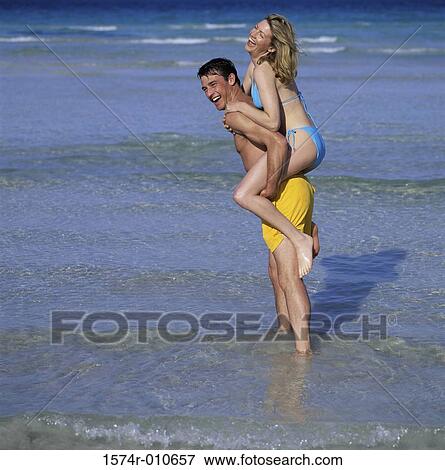 Stock Photo - Side profile of a young woman riding piggyback on a young man at the beach. Fotosearch