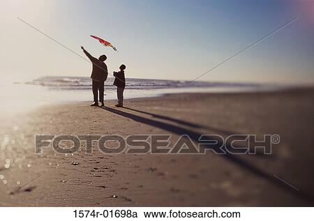 Stock Image - Silhouette of a father and son flying a kite on the beach. Fotosearch