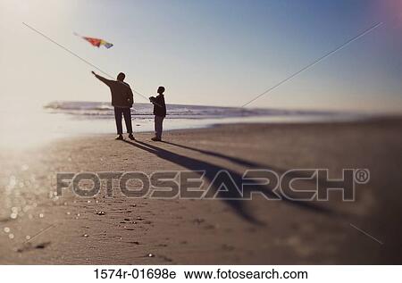 Stock Image - Silhouette of a father and son flying a kite on the beach. Fotosearch