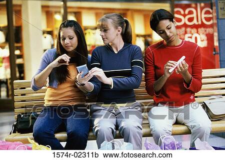 Stock Image - Three teenage girls sitting on a bench in a shopping mall applying make-up. Fotosearch