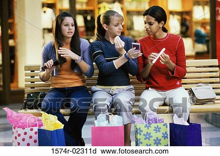 Stock Image - Three teenage girls sitting on a bench in a shopping mall applying make-up. Fotosearch