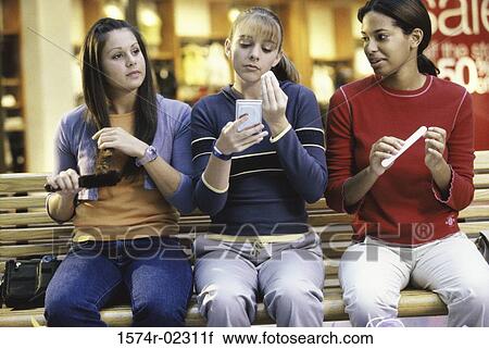 Stock Image - Three teenage girls sitting on a bench in a shopping mall applying make-up. Fotosearch
