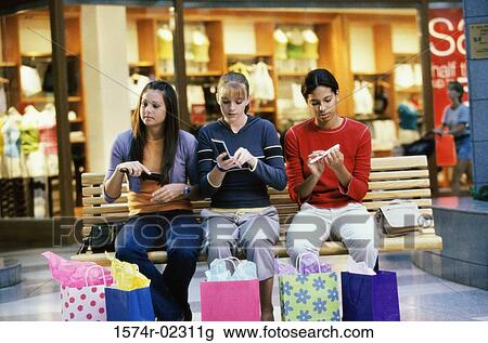 Stock Image - Three teenage girls sitting on a bench in a shopping mall applying make-up. Fotosearch