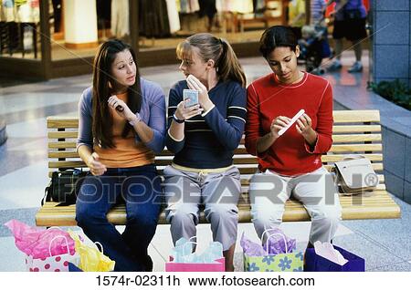 Stock Image - Three teenage girls sitting on a bench in a shopping mall applying make-up. Fotosearch