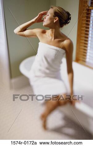 Stock Image - Young woman sitting in a towel on the edge of a bathtub. Fotosearch