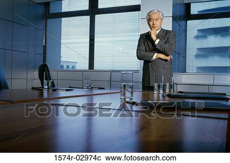 Stock Image - Businessman standing in a boardroom with his hand on his chin. Fotosearch