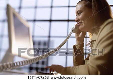 Stock Image - Businesswoman talking on a telephone sitting in front of a computer monitor. Fotosearch