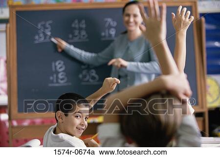 Stock Image - Female teacher pointing to a chalkboard in a classroom with her students raising their hands. Fotosearch