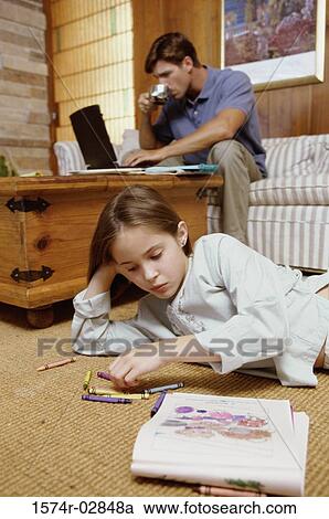 Stock Image - Girl lying on the floor with her father working on a laptop behind her. Fotosearch