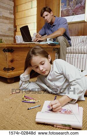 Stock Image - Girl lying on the floor with her father working on a laptop behind her. Fotosearch