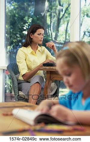 Stock Image - Girl writing in a book with her mother sitting behind her. Fotosearch