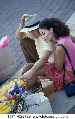 Stock Image - High angle view of a young couple sitting on the sidewalk with shopping bags. Fotosearch