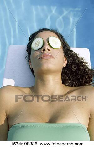 Stock Photograph - High angle view of a young woman lying near a swimming pool with cucumber slices on her eyes. Fotosearch
