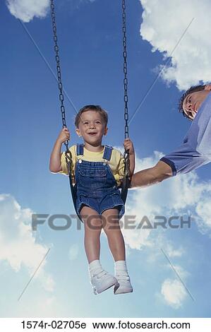 Stock Image - Low angle view of a father pushing his son on a swing. Fotosearch