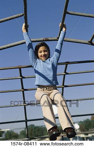 Stock Image - Low angle view of a girl swinging on monkey bars at a playground. Fotosearch
