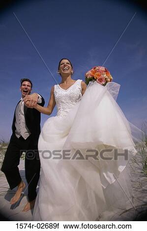 Stock Image - Low angle view of a newlywed couple running on sand. Fotosearch