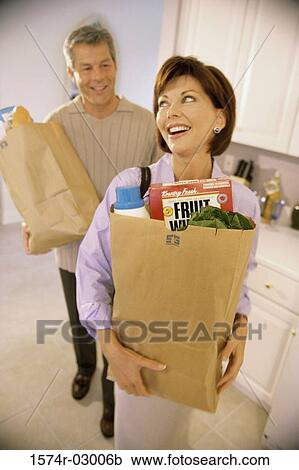 Stock Image - Mid adult couple standing in a kitchen holding grocery bags. Fotosearch