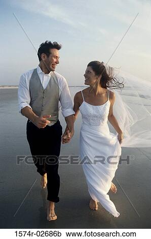 Stock Image - Newlywed couple holding hands running on the beach. Fotosearch