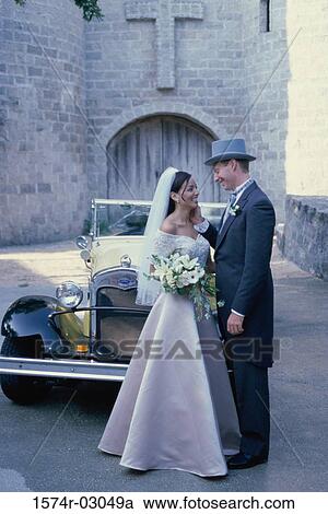 Stock Image - Newlywed couple standing in front of an antique car. Fotosearch