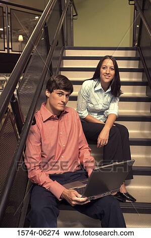 Picture - Portrait of a businessman and a businesswoman sitting on the stairs with a laptop. Fotosearch