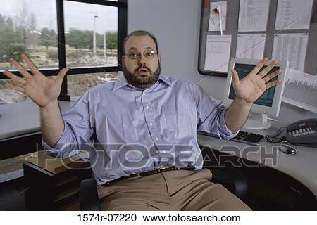 Stock Image - Portrait of a businessman seated at an office desk with his hands raised. Fotosearch
