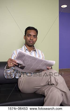 Stock Image - Portrait of a businessman sitting on a couch reading a newspaper in an office. Fotosearch