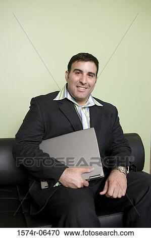 Portrait of a businessman sitting on a couch in an office holding a laptop View Large Photo Image Stock Image - Portrait of a businessman sitting on a couch in an office holding a laptop. Fotosearch