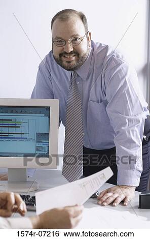 Stock Photograph - Portrait of a businessman standing leaning against a table in an office. Fotosearch