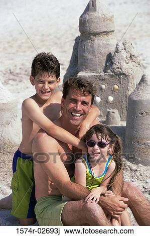 Stock Image - Portrait of a father with his son and daughter sitting on the beach. Fotosearch
