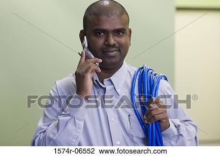 Stock Image - Portrait of a technician carrying computer cables on his shoulder talking on a mobile phone. Fotosearch
