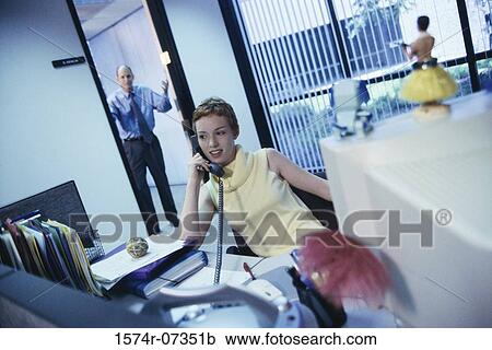 Stock Image - Secretary talking on a telephone with a businessman standing in a doorway behind. Fotosearch