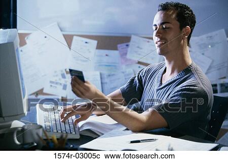 Stock Image - Side profile of a young man working on a computer holding a credit card. Fotosearch