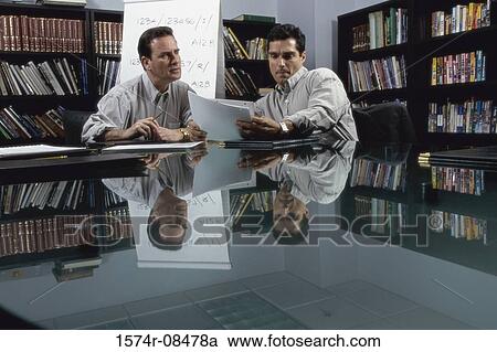 Stock Image - Two businessmen seated at a table reading documents. Fotosearch