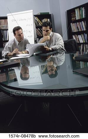 Stock Image - Two businessmen seated at a table reading documents. Fotosearch