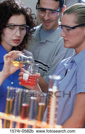 Stock Image - Two young women and a young man pouring liquid from a flask into a measuring beaker. Fotosearch
