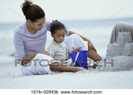 Stock Image - Woman sitting on a beach with her daughter building a sandcastle. Fotosearch