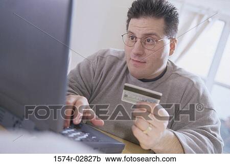 Stock Image - Young man sitting in front of a laptop holding a credit card. Fotosearch