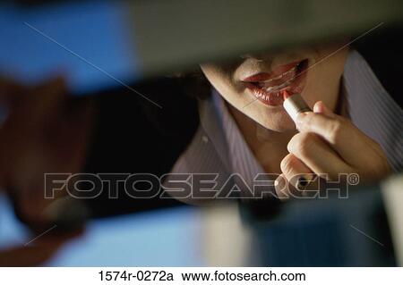 Stock Image - Young woman applying lipstick in a car rear view mirror. Fotosearch