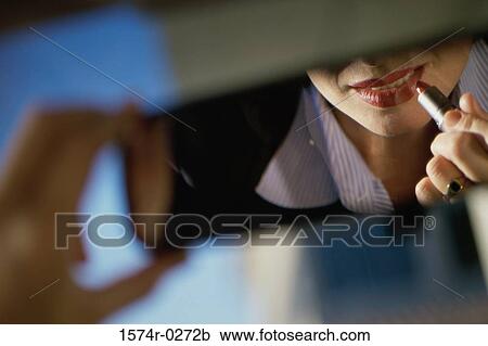 Stock Image - Young woman applying lipstick in a car rear view mirror. Fotosearch